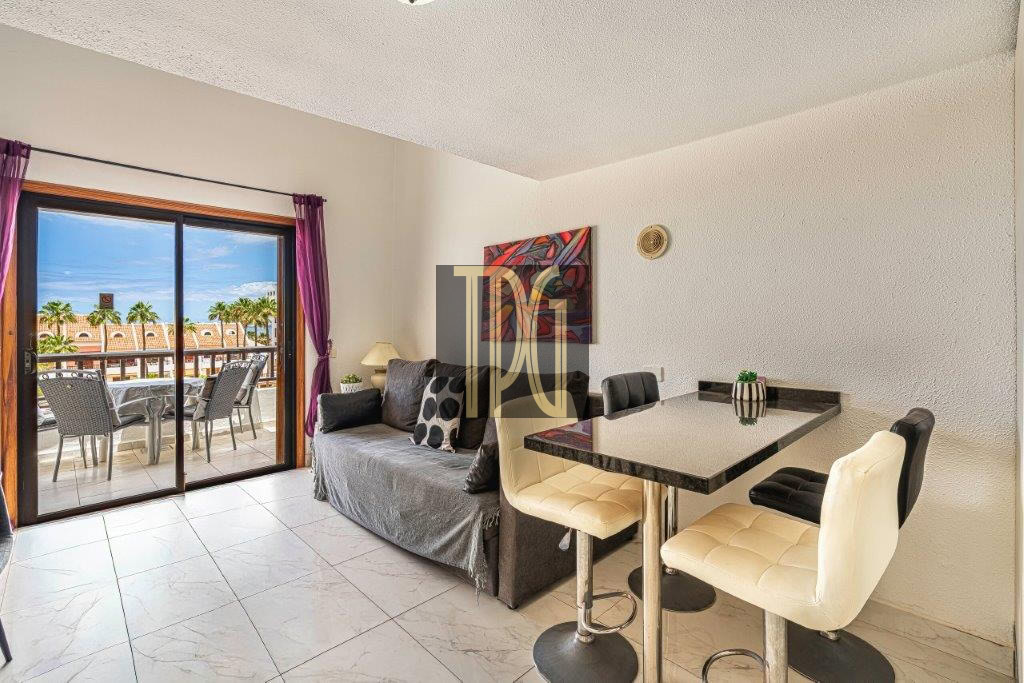 Living area with a gray sofa, bar-height dining table and cream stools, and a sliding glass door to a balcony with outdoor furniture and palm trees outside.