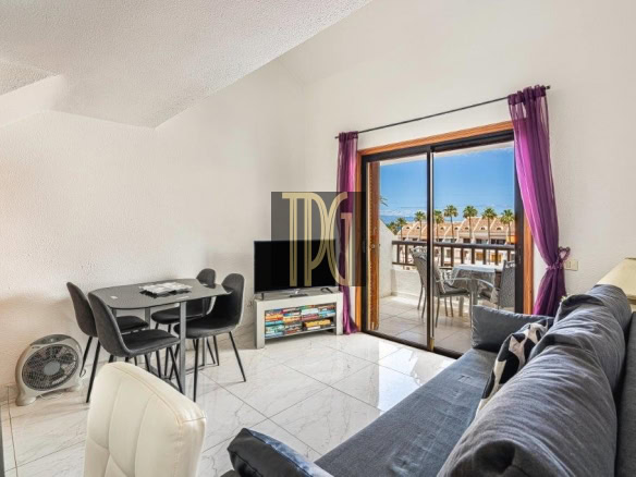 Bright living room with a gray sectional, dining table for four, and a TV stand; sliding doors open to a balcony with purple curtains and an ocean view.