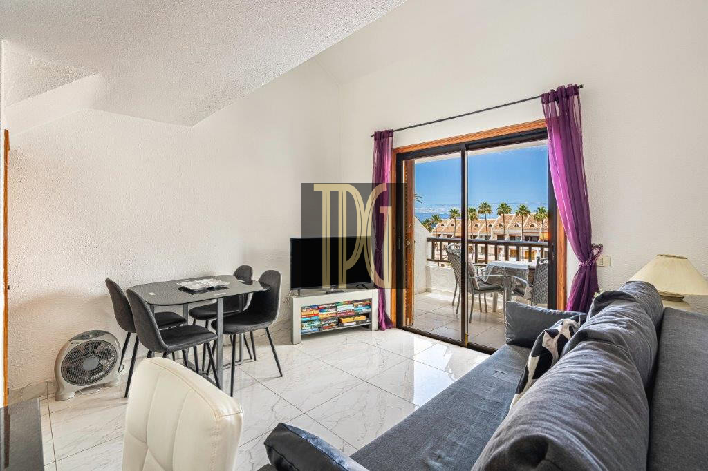 Bright living room with a gray sectional, dining table for four, and a TV stand; sliding doors open to a balcony with purple curtains and an ocean view.