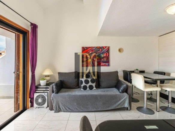 Living room with a gray sofa, purple curtains, and a glass sliding door to a patio on the left; a small lamp and plant on a side table beside the sofa; a colorful wall art piece above the couch; dining table with cream chairs and a kitchen area in the background.