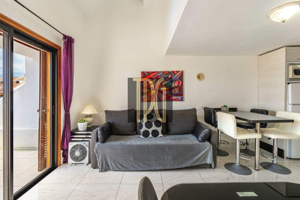 Living room with a gray sofa, purple curtains, and a glass sliding door to a patio on the left; a small lamp and plant on a side table beside the sofa; a colorful wall art piece above the couch; dining table with cream chairs and a kitchen area in the background.