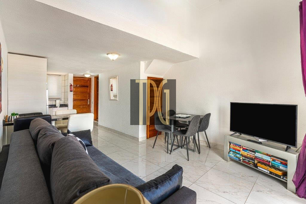 Open-plan living room with a dark gray sectional facing a flat-screen TV on a low stand and a compact bookshelf below.