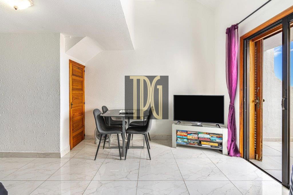 Modern living area with a glass dining table and four black chairs, a TV on a low stand, and a bookshelf full of books, beside open sliding glass doors with purple curtains.