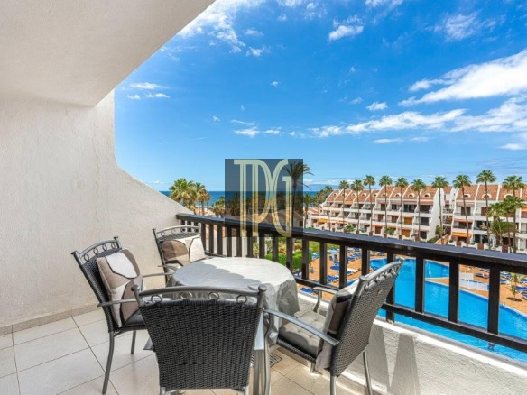 Balcony with a round table and four wicker chairs overlooking a resort pool, palm trees, and the sea on the horizon.