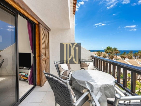 Balcony with a round table covered by a cloth and wicker chairs, overlooking a beach and palm trees under a blue sky.