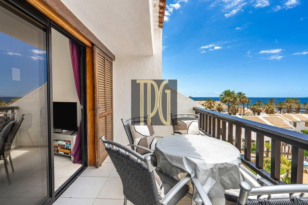 Balcony with a round table covered by a cloth and wicker chairs, overlooking a beach and palm trees under a blue sky.