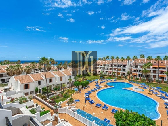 Resort swimming pool surrounded by blue lounge chairs, white buildings with terracotta roofs, and palm trees under a bright blue sky with the ocean in the distance.