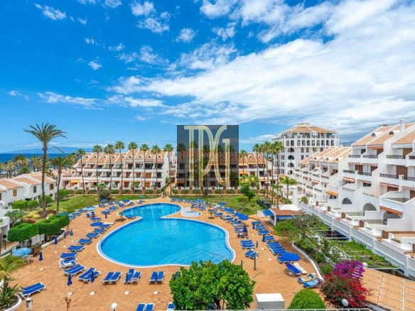 Resort pool area with a curved blue pool, rows of blue loungers, palm trees, and white villa-style buildings.