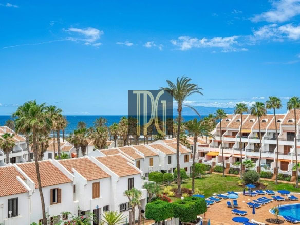 Seaside resort with white buildings, orange-tiled roofs, palm trees, and a blue pool surrounded by lounge chairs.