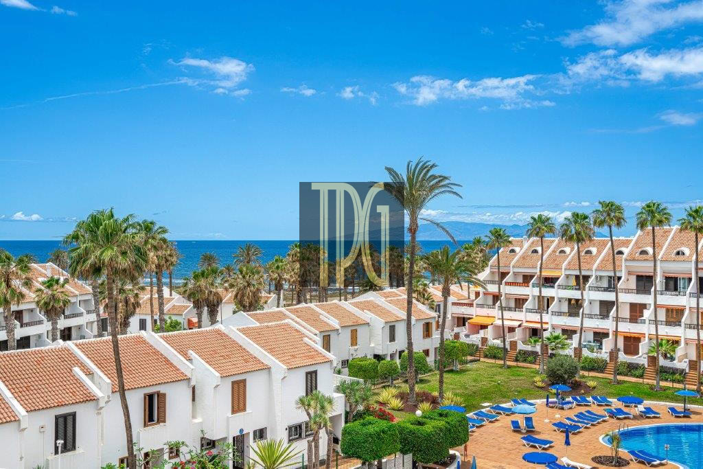Seaside resort with white buildings, orange-tiled roofs, palm trees, and a blue pool surrounded by lounge chairs.