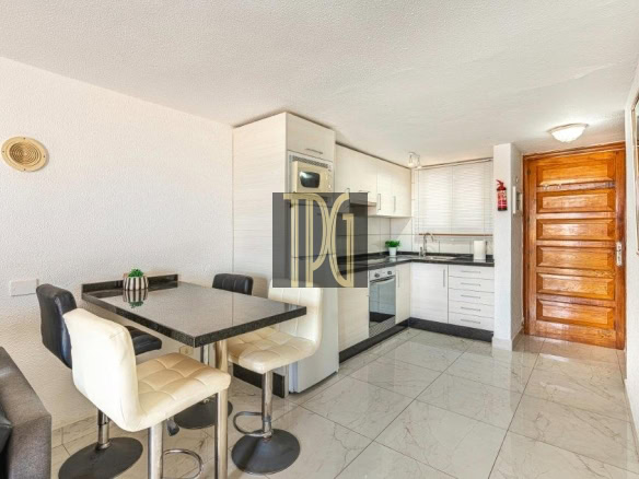 Compact modern kitchen with white cabinets, black countertops, and a small dining table with four cushioned stools near a living area sunlit by natural light.