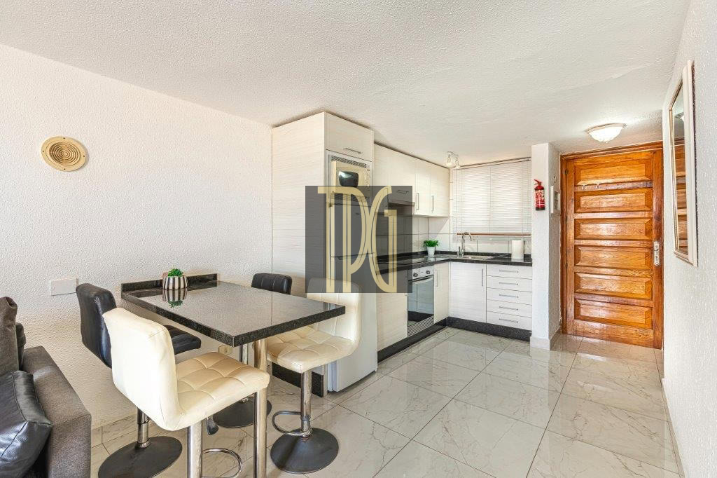 Compact modern kitchen with white cabinets, black countertops, and a small dining table with four cushioned stools near a living area sunlit by natural light.