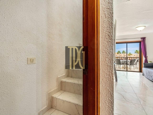 Doorway opens into a bright living area; stairs on the left and a balcony with outdoor dining beyond the glass doors showing palm trees outside.