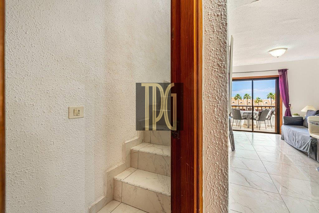 Doorway opens into a bright living area; stairs on the left and a balcony with outdoor dining beyond the glass doors showing palm trees outside.