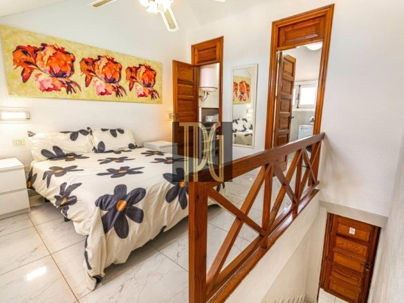 Bright bedroom featuring a queen bed with a flower-pattern duvet, a white nightstand, and a wooden railing by the stairs.