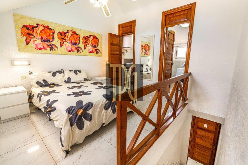 Bright bedroom featuring a queen bed with a flower-pattern duvet, a white nightstand, and a wooden railing by the stairs.