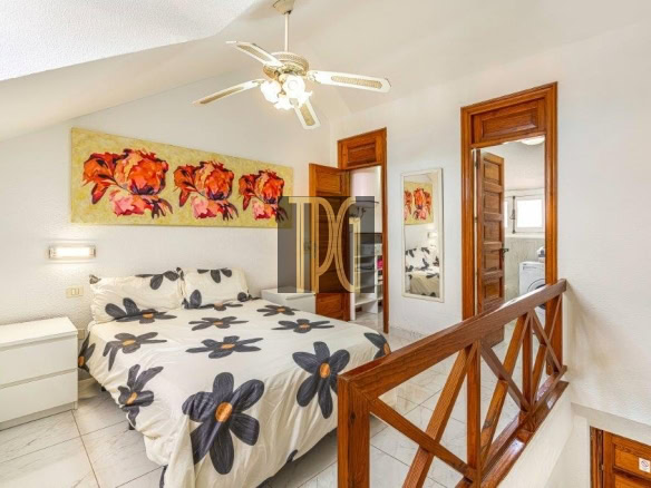 Queen bed with white bedding featuring large black flowers with orange centers, white nightstand, ceiling fan, and wooden railing in a bright bedroom.