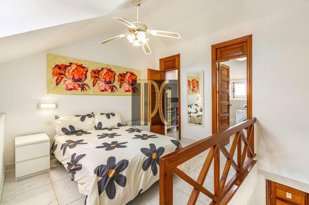 Queen bed with white bedding featuring large black flowers with orange centers, white nightstand, ceiling fan, and wooden railing in a bright bedroom.