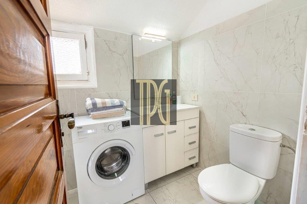 Small bathroom with a front-loading washing machine under a stack of towels, white vanity, and toilet.