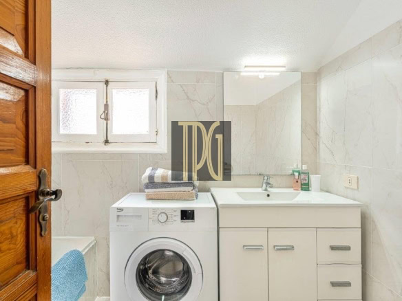 Small bathroom with a front-loading washing machine beside a white vanity, mirror, and window on tiled walls.