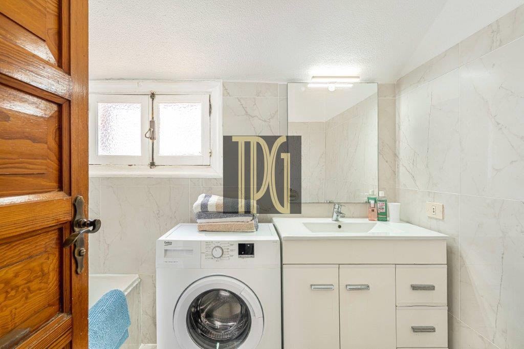 Small bathroom with a front-loading washing machine beside a white vanity, mirror, and window on tiled walls.