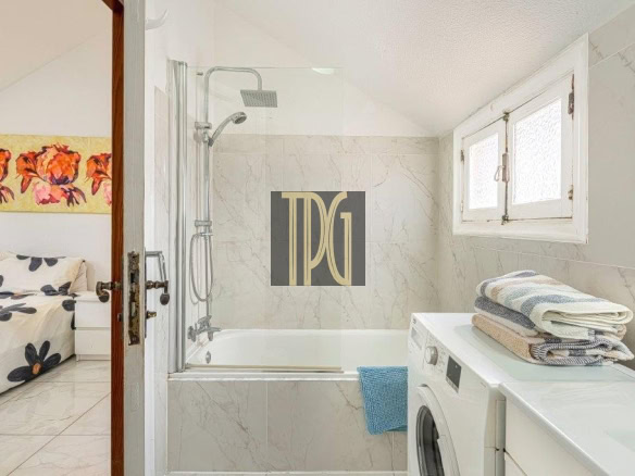 Bathroom with a glass shower enclosure over a tub, marble walls, and a front-loading washing machine with folded towels on top; door opens to a bedroom with floral artwork visible.