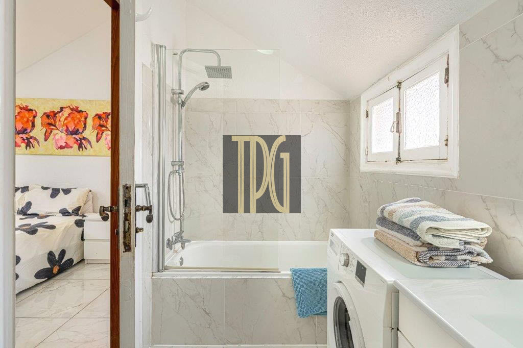 Bathroom with a glass shower enclosure over a tub, marble walls, and a front-loading washing machine with folded towels on top; door opens to a bedroom with floral artwork visible.