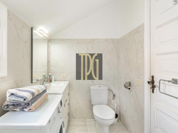 Small marble-tiled bathroom with a front-loading washing machine under a counter, folded towels on top, sink with mirror, and a toilet against the wall.