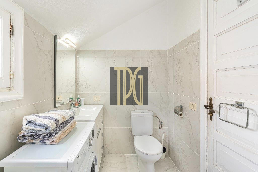 Small marble-tiled bathroom with a front-loading washing machine under a counter, folded towels on top, sink with mirror, and a toilet against the wall.