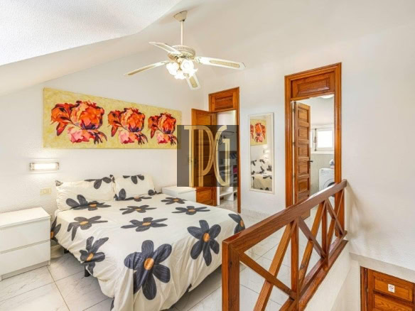 Bright upstairs bedroom with a queen bed in white bedding patterned with navy flowers, a white nightstand, and a colorful wall art piece above the bed.