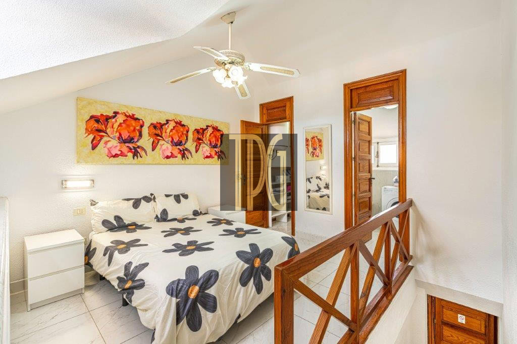 Bright upstairs bedroom with a queen bed in white bedding patterned with navy flowers, a white nightstand, and a colorful wall art piece above the bed.