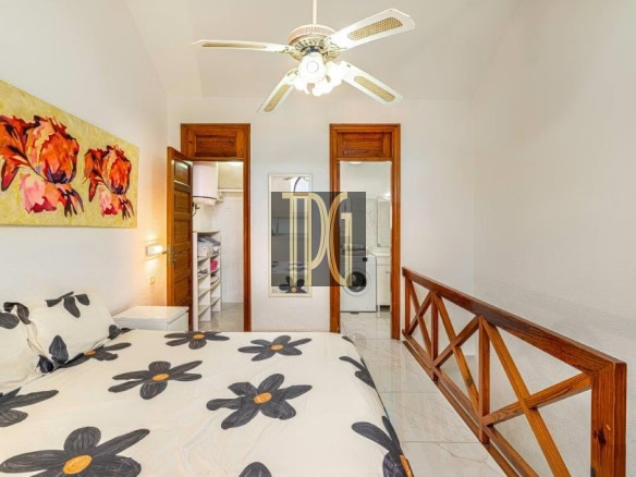 Bright bedroom with a bed covered in a black-and-yellow floral duvet, wall art of red flowers, a ceiling fan, and wooden railing along the right. Two doors in back lead to a laundry area.