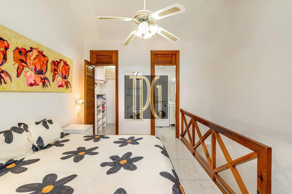 Bright bedroom with a bed covered in a black-and-yellow floral duvet, wall art of red flowers, a ceiling fan, and wooden railing along the right. Two doors in back lead to a laundry area.