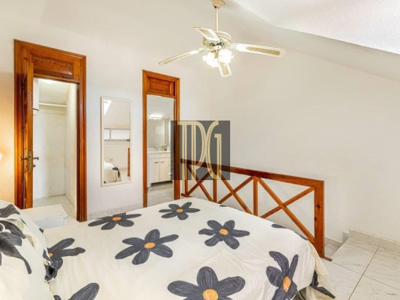 Bedroom with white walls, wooden headboard, floral duvet, ceiling fan, full-length mirror, and two doorways leading to a bathroom and kitchenette.