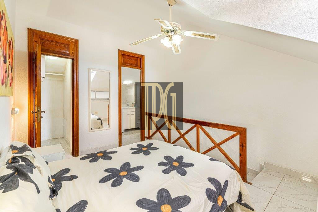 Bedroom with white walls, wooden headboard, floral duvet, ceiling fan, full-length mirror, and two doorways leading to a bathroom and kitchenette.