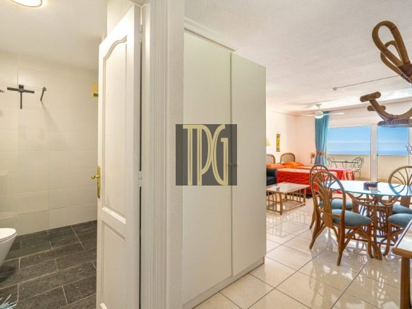 Entry view showing a bathroom on the left, with shower and toilet, as a white cabinet sits in a bright hallway leading to a sunny living/dining area with a balcony and ocean view.