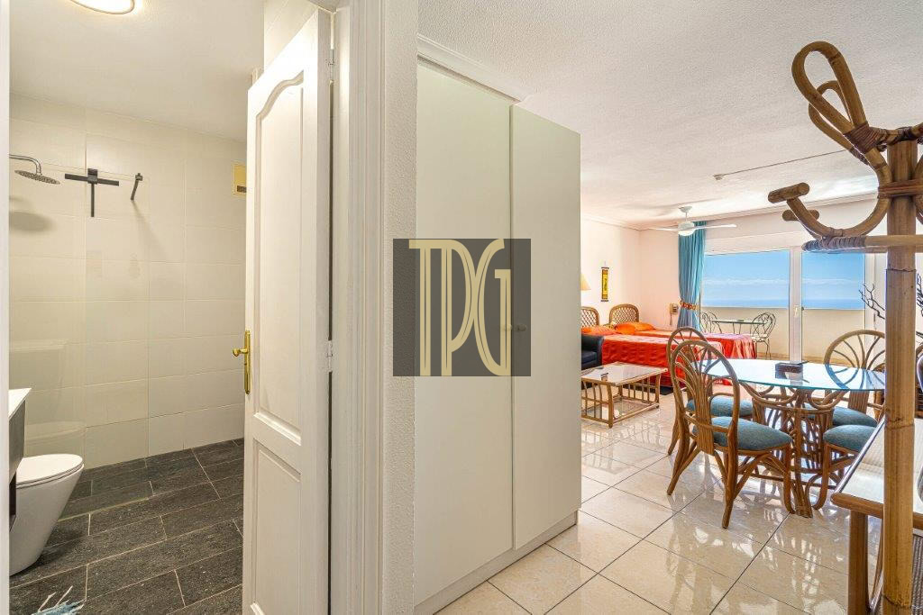 Entry view showing a bathroom on the left, with shower and toilet, as a white cabinet sits in a bright hallway leading to a sunny living/dining area with a balcony and ocean view.