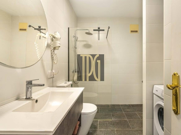 Modern bathroom with white double vanity, round mirror, wall-mounted hair dryer, toilet and glass-enclosed shower. A front-loading washing machine sits near the door.