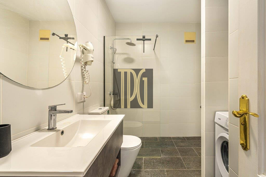 Modern bathroom with white double vanity, round mirror, wall-mounted hair dryer, toilet and glass-enclosed shower. A front-loading washing machine sits near the door.