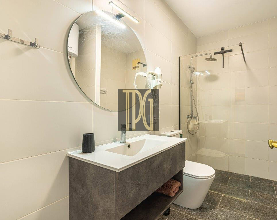 Modern bathroom with a round mirror above a white sink and dark vanity, glass shower enclosure, and toilet.