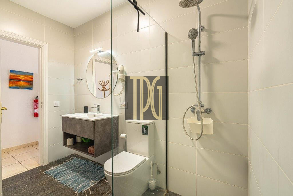 Modern bathroom with a glass shower enclosure, wall-mounted sink, and toilet; blue rug on a dark floor.