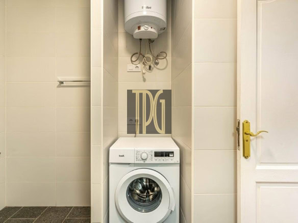 White front-loading washing machine under a wall-mounted water heater in a small tiled laundry alcove, with a white door to the right and brass handle.