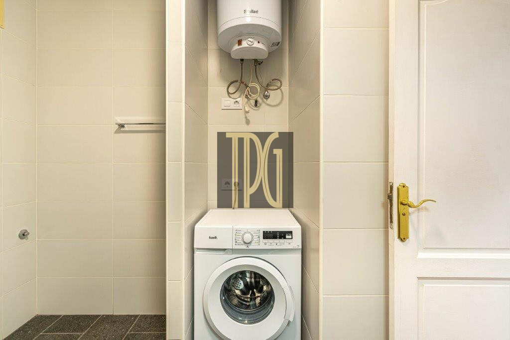 White front-loading washing machine under a wall-mounted water heater in a small tiled laundry alcove, with a white door to the right and brass handle.