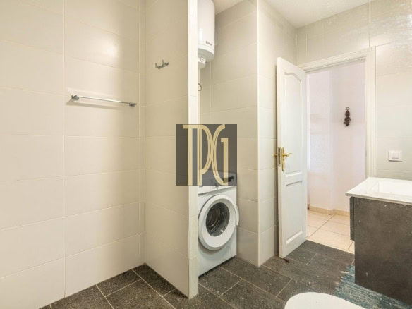 Bathroom with a front-loading washing machine tucked into an alcove beside an open door; beige tiled walls and dark flooring.
