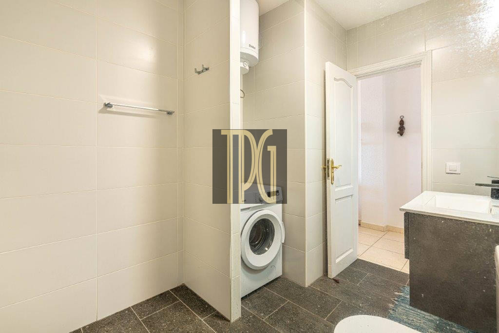 Bathroom with a front-loading washing machine tucked into an alcove beside an open door; beige tiled walls and dark flooring.