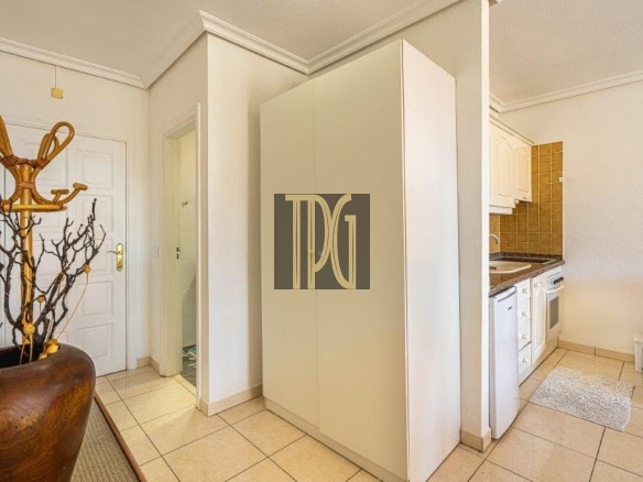 Bright kitchen area with white cabinets and beige tiled floor; a tall white wardrobe centered, a wooden coat rack in a large pot to the left, and a small kitchen with brown tile backsplash on the right.
