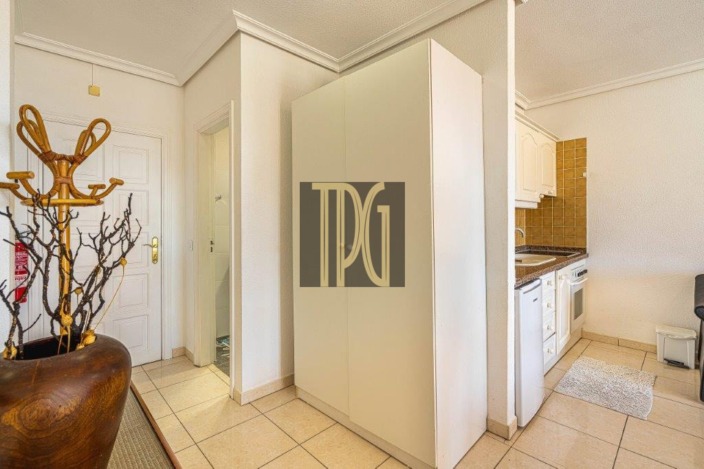 Bright kitchen area with white cabinets and beige tiled floor; a tall white wardrobe centered, a wooden coat rack in a large pot to the left, and a small kitchen with brown tile backsplash on the right.