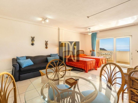 Living area and bedroom in a seaside suite: glass dining table, wicker chairs, black sofa with blue cushions, two red-bedspreads, and a balcony with ocean view on the right.