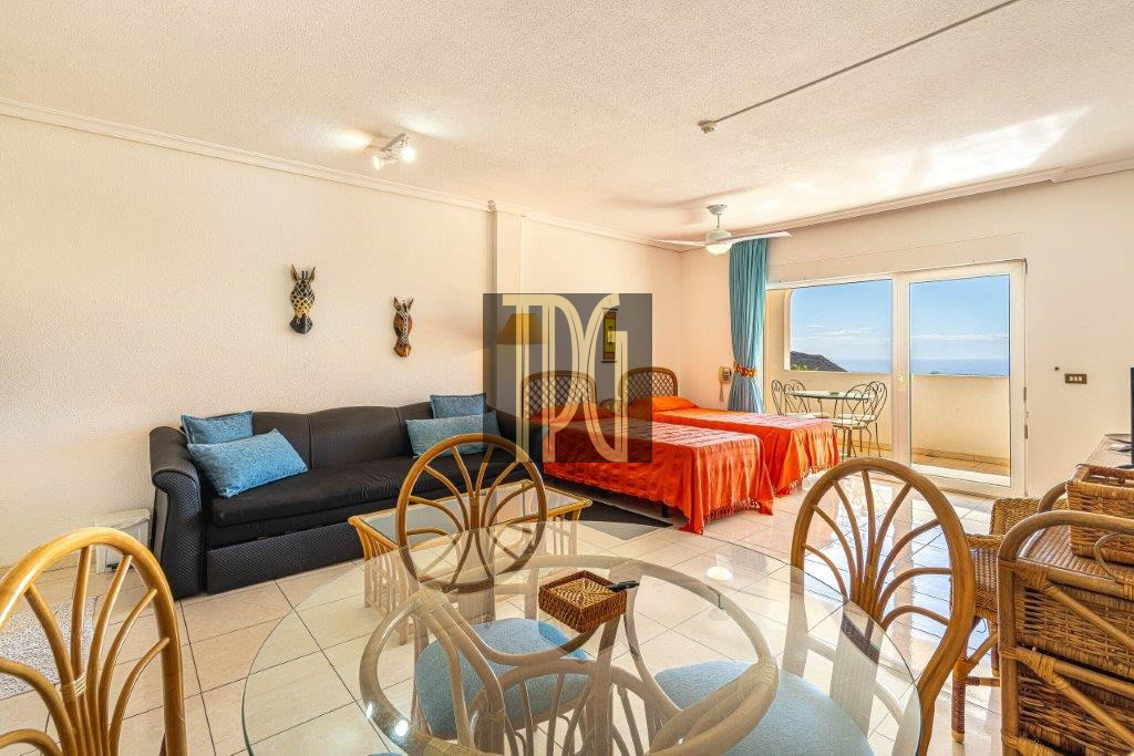 Living area and bedroom in a seaside suite: glass dining table, wicker chairs, black sofa with blue cushions, two red-bedspreads, and a balcony with ocean view on the right.