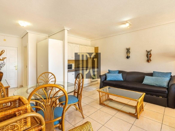Living room with a round glass dining table and wicker chairs, adjacent to a dark sofa with blue pillows on a tiled floor.